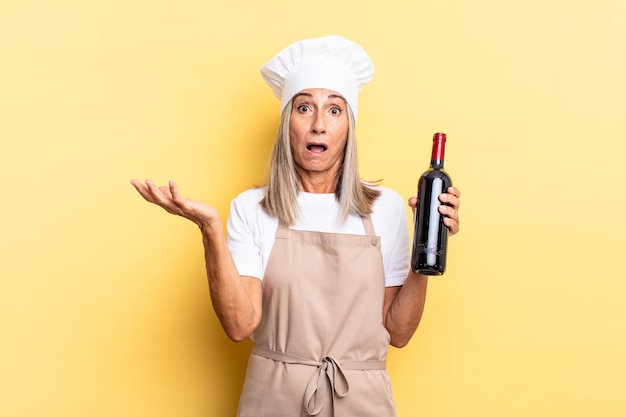 Chef-kokvrouw van middelbare leeftijd die zich extreem geschokt en verrast, angstig en in paniek voelt, met een gestrest en geschokte blik met een wijnfles