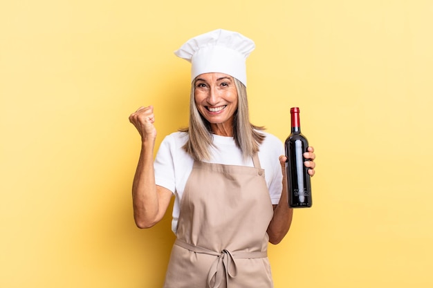 Chef-kokvrouw van middelbare leeftijd die zich geschokt, opgewonden en gelukkig voelt, lacht en succes viert, en zegt wauw! een wijnfles vasthouden