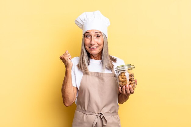 Chef-kokvrouw van middelbare leeftijd die zich geschokt, opgewonden en gelukkig voelt, lacht en succes viert, en zegt wauw! met koekjes