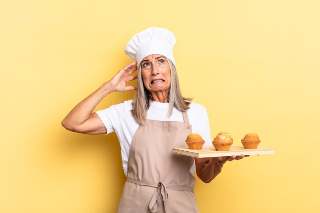 Chef-kokvrouw van middelbare leeftijd die zich gestrest, bezorgd, angstig of bang voelt, met handen op het hoofd, in paniek bij vergissing en met een dienblad met muffins