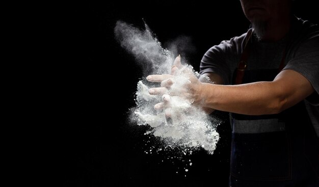 베이커리 음식을 요리하기 위해  밀가루 먼지를 준비하는 요리사 노인 요리사 손을 박수 는  가루 먼지가 공중에서 날아갑니다 고속 셔터와 함께 공중에서 움직임을 멈추는 밀가루 검은 배경