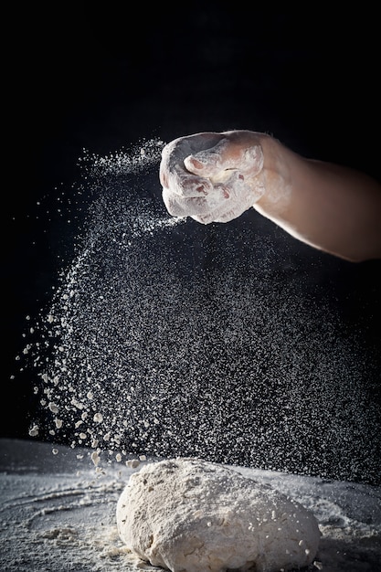 Chef prepares the dough with flour. male sprinkling flour over dough on table on dark background. Vertical. Copy space. Conceptn of flying food. Gluten free dough for pasta, bakery or pizza