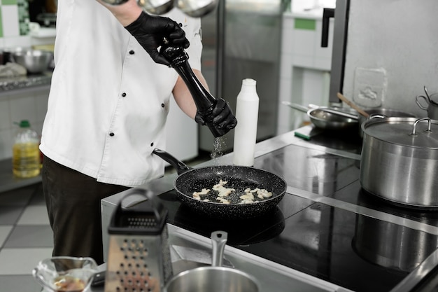 Chef's hands in a tunic that adds salt or pepper to a dish when frying in a pan