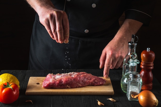 Chef sprinkles the raw beef meat with salt. Preparing meat before baking. Working environment in the kitchen of restaurant or cafe