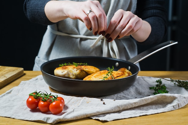 Chef sprinkles thyme on chicken Breasts in a frying pan. 