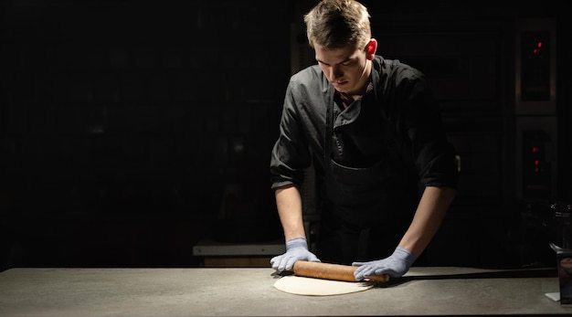 A chef in a suit rolls out thin pizza dough on a table in a\
pizzeria next to a place for text. banner and dark background.