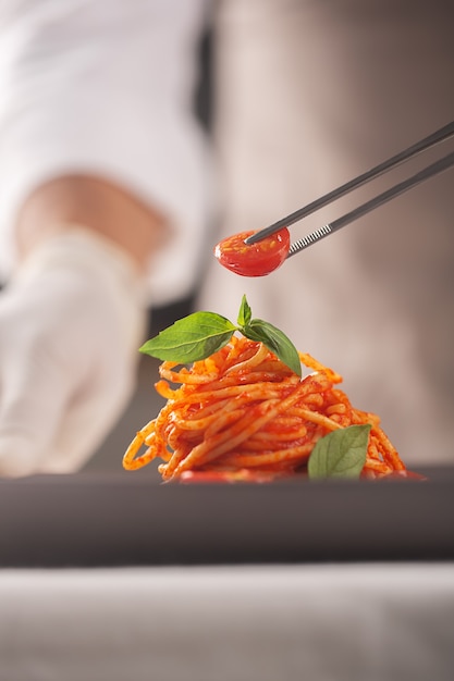 A chef in a white uniform and gloves serves cherry tomatoes in pasta in tomato sauce with basil leaves with tweezers . Haute cuisine. Plating food