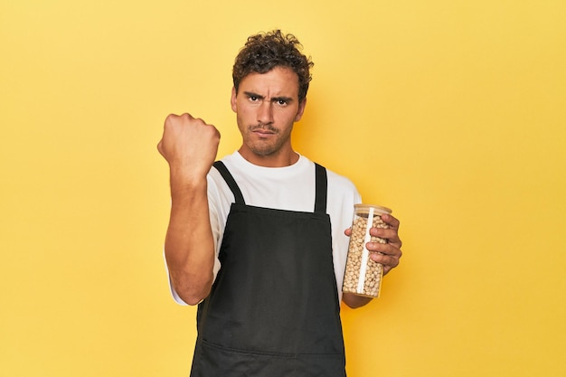 Chef with chickpeas jar on yellow showing fist to camera aggressive facial expression
