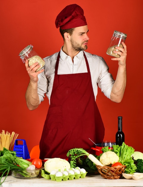 Chef with curious face holds jars with porridge on red background. Man in cook hat and apron uses groats. Cook works in kitchen near table with vegetables and tools. Cuisine and cooking concept.