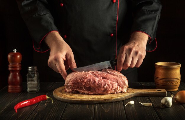 The chef with a knife in his hand in the kitchen prepares to cut raw meat before baking or barbecue