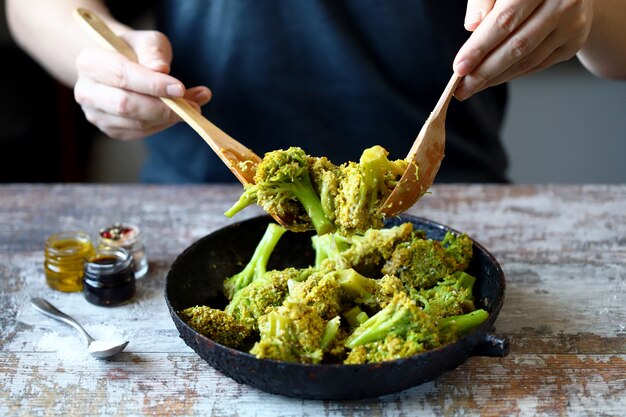 A chef with a wooden spatula and spoon mixes broccoli in a pan.