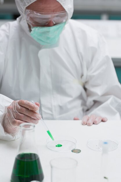 Chemist in protective suit adding green liquid to petri dish