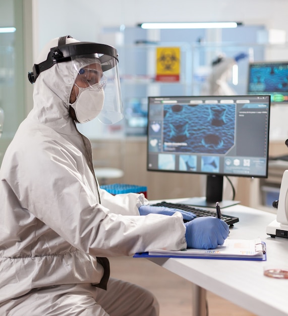 Chemist researcher dressed in ppe suit with visor writing on clipboard. Examining vaccine evolution using high tech technology and chemistry tools for scientific research virus development.