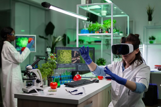 Женщина-врач ученый-химик в наушниках виртуальной реальности анализирует микробиологический эксперимент, работающий в лаборатории фармацевтической больницы. Генетически модифицированные растения экрана компьютера