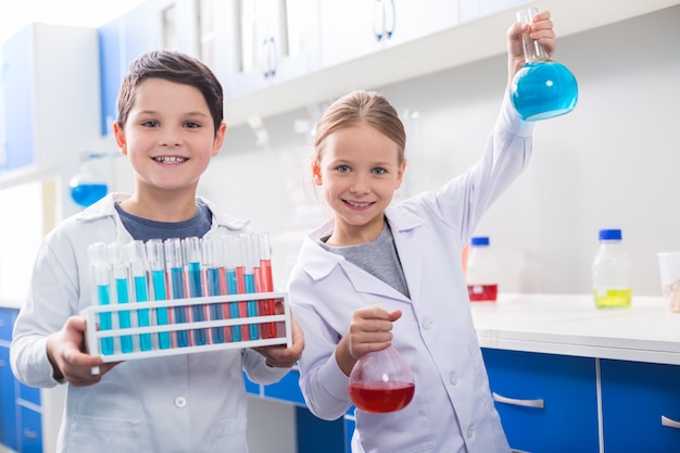 Lezione di chimica. simpatici bambini intelligenti positivi che ti guardano e trasportano reagenti chimici mentre hanno una lezione di chimica