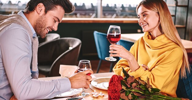 Cherful praten in restaurant. Paar met rode wijn in glazen in restaurant. Rode rozen en kaarsen liggen op tafel bij de rode wijn. Jonge vrouw in mosterdtrui kijkt naar haar vriendje