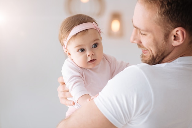 一瞬一瞬を大切に。家に立って優しい感情を表現しながら幼児の女の子を抱き締める陽気な笑顔のひげを生やした父親