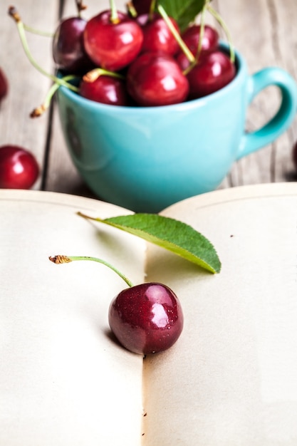 Cherry in the beautiful turquoise cup and old book on a wooden table.