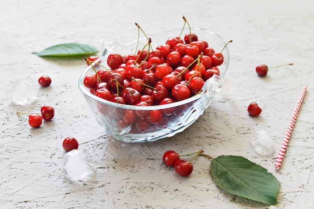 Cherry berries with leaves in a glass bowl and ice cubes on a gray background. Side view