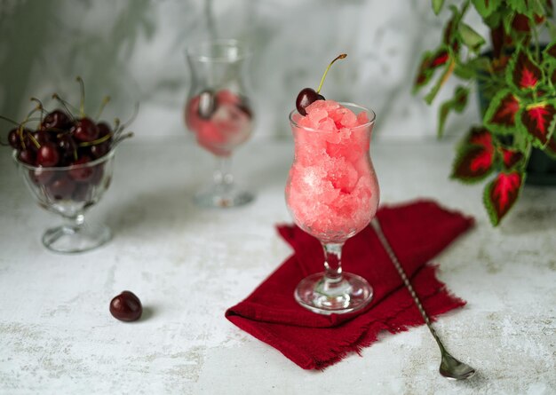 Granita di ciliegie, frutti di bosco in vaso di vetro e bevanda rinfrescante con ghiaccio su fondo grigio chiaro