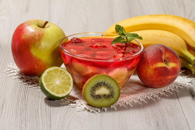 Cherry jelly with strawberry pieces in the glass bowl topped mint leaves, nectarine, kiwi, lime, apple and banana on the white napkin