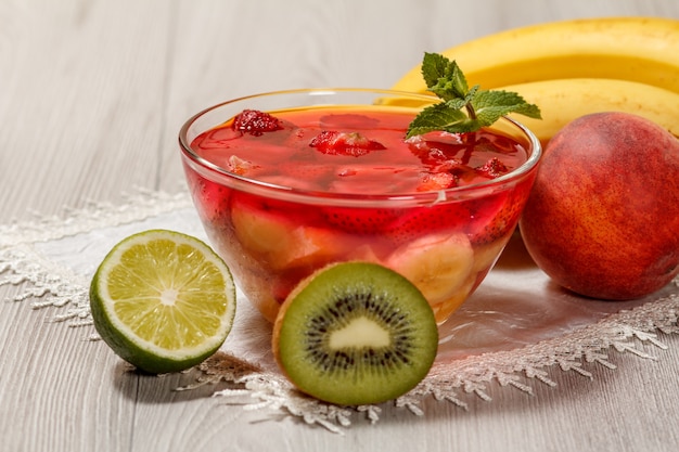 Cherry jelly with strawberry pieces in the glass bowl topped mint leaves, nectarine, kiwi, lime and banana on the white napkin