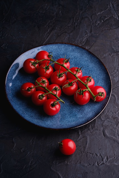 Cherry tomato branch, fresh ripe organic vegetables in blue plate on dark black surface, angle view