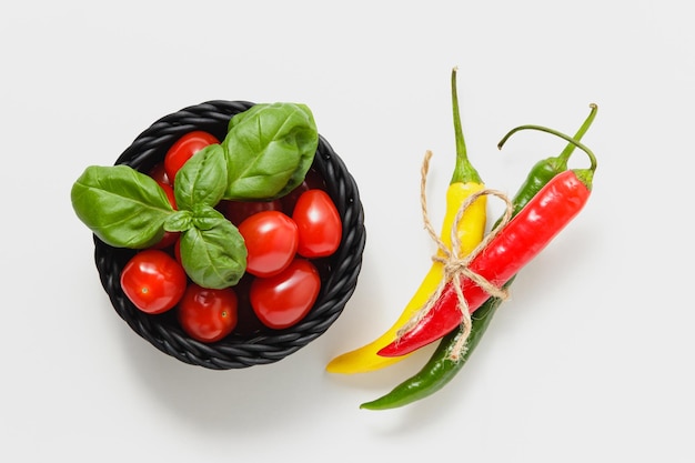 Cherry tomatoes in black basket and green basil leaves on white background