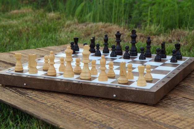 Chess board with chess pieces on wooden desk with green grass on the background. Selective focus on white pieces. Outdoors chess game.