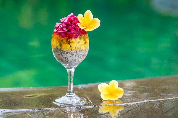スイミングプールの水のクローズアップの背景に朝食用のグラスに赤いドラゴンフルーツパッションフルーツマンゴーとアボカドとチアシードプリン健康的な食事の概念