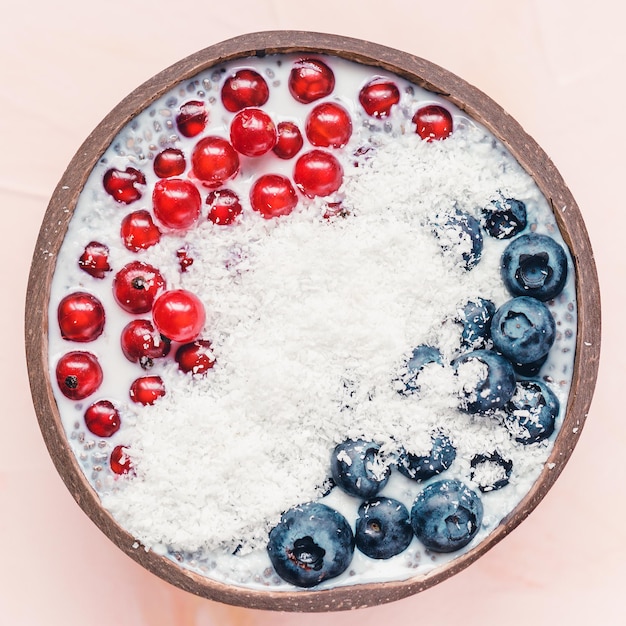 Chia-zaadpudding 's nachts met verse bosbessen, rode aalbessen en kokosvlokken in een kokoskom Close-up bovenaanzicht plat leggen Superfood en veganistisch voedselconcept Roze achtergrond