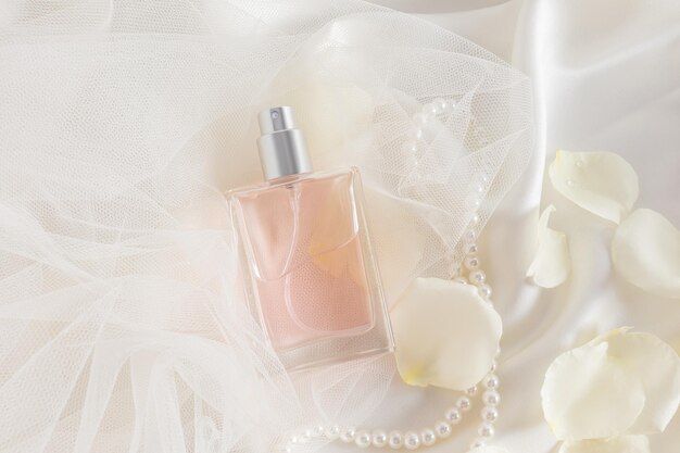 A chic bottle of women's perfume or eau de parfum with a delicate smell of roses lies on a satin fabric of the color of cream and rose petals
