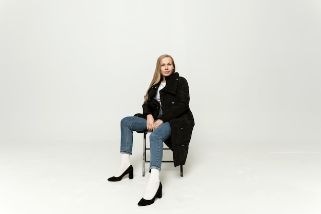 Chic fashion model in full growth sitting on a chair on a white background.blonde of European appearance on a white background in high heels and in a jacket