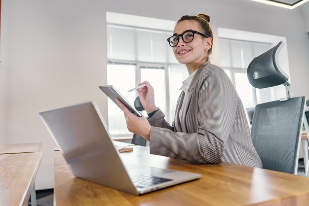 A chic professional woman with a tablet and stylus gives a pleased smile poised at her desk