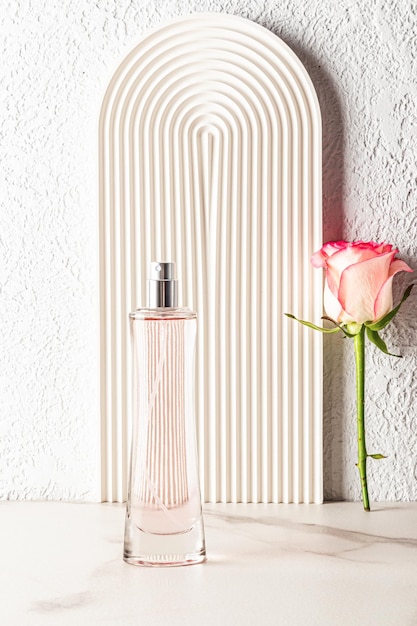 A chic tall bottle of women's perfume against the background of a white decorative arch with a rose flower Vertical view empty bottle