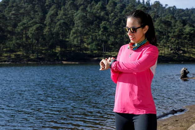 Chica revisando su reloj inteligente a la orilla de un lago despues de correr