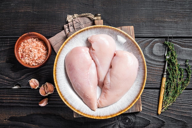 Chicken breast fillet raw meat in plate with herbs. Black Wooden background. Top view.