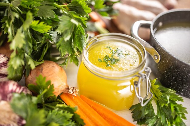 Chicken broth in a glass jar raw chicken legs carrot onion celery herbs garlic and fresh vegetables.