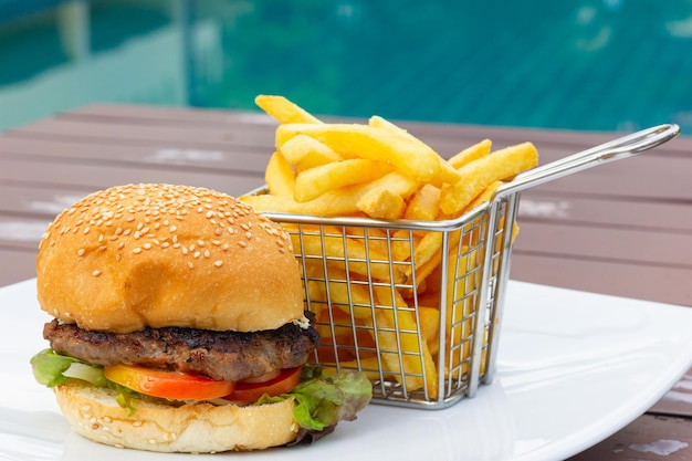 Chicken burger with stainless steel basket of french fries on white plate by swimming pool