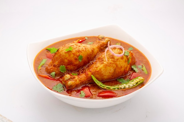 Chicken curry or masala , spicy reddish chicken leg piece dish garnished with coriander leaf and  fresh green chilli arranged in a white ceramic bowl with white background,isolated.