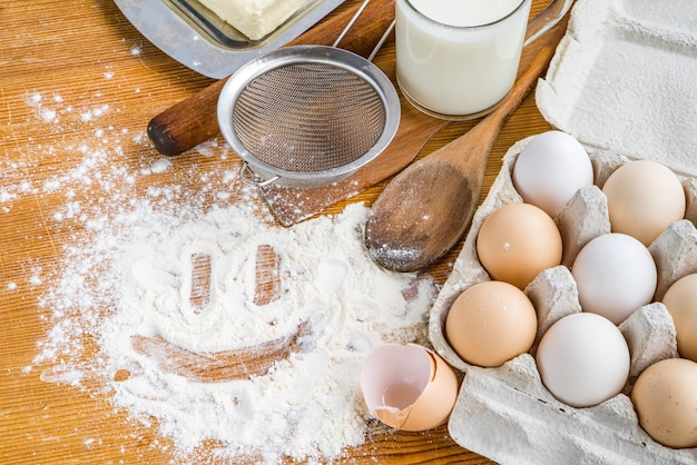 Куриные яйца в картонном подносе рядом с кухонной утварью и разбросанная белая мука на столе и сердце, нарисованное на муке Кулинария Рецепты кулинарии