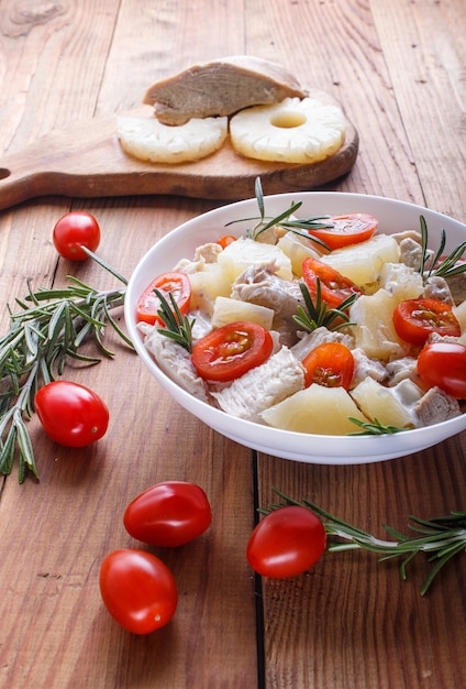 Chicken fillet salad with rosemary, pineapple and cherry tomatoes on brown wooden background.