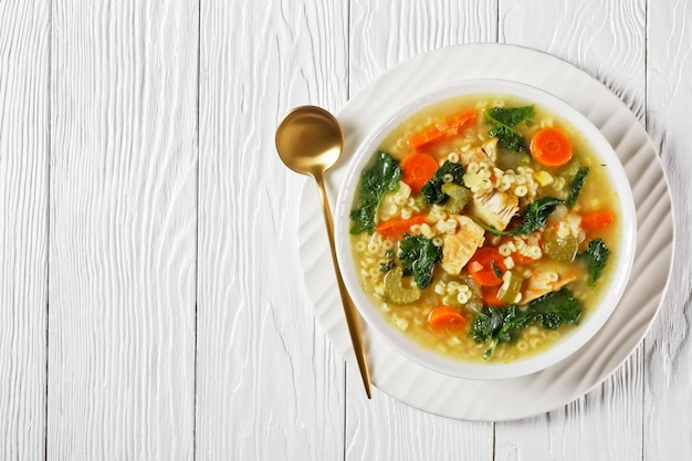 Chicken kale soup with pasta anellini and vegetables in a white bowl with golden spoon on a wooden table, flat lay, free space, italian cuisine