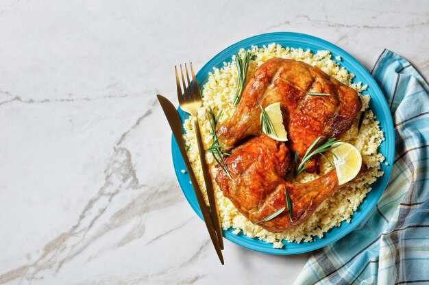 Chicken leg quarters served over the millet on a plate with golden cutlery, lemon wedges, and fresh rosemary sprigs on a white marble stone background, top view, close-up, copy space