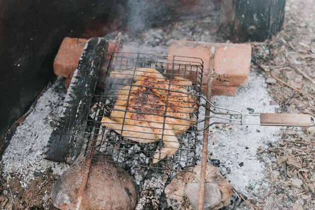 Chicken meat food are fried on grill on smoldering coals or ember from a campfire on the ground on summer or autumn day barbecue in camping conditions survival on countryside and wild rustic life