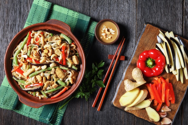 Chicken satay rice noodle with green beans, pepper and eggplant in a bowl on a wooden table with ingredients, horizontal view from above, flat lay