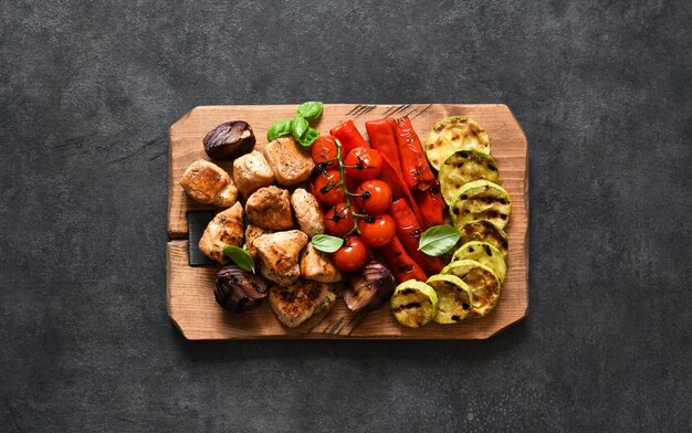 Chicken skewers with grilled vegetables on a wooden board on a black surface