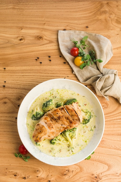 Chicken steak with broccoli in cream sauce in a plate on a light wooden surface top view.