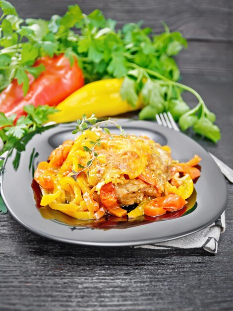 Chicken stewed with tomatoes, yellow and red bell peppers and cheese in a plate on napkin, thyme, parsley and garlic on black wooden board background