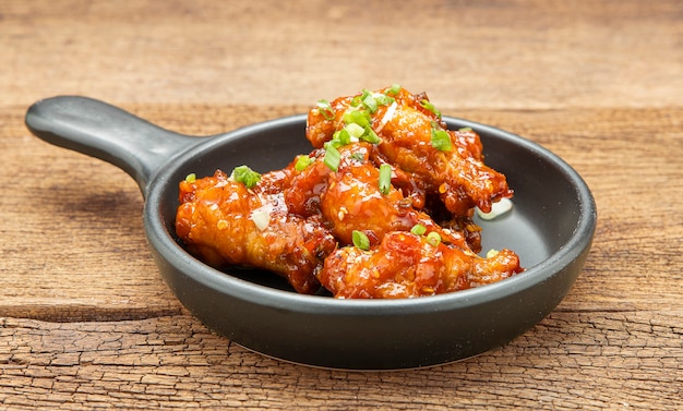 Chicken stir-fried with spicy sauce sprinkled with white sesame and chopped scallions is placed in a black ceramic pan on a wooden table.